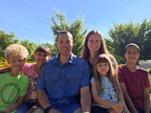 Family Photo Outdoors A family of six sitting together outdoors on a sunny day with trees and flowers in the background.